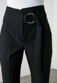 Černé společenské kalhoty s širok ým pasem, vybavené velkou stříbrnou kruhovou sponou a vázaným textilním páskem. Hladká textura a přiléhavý střih.