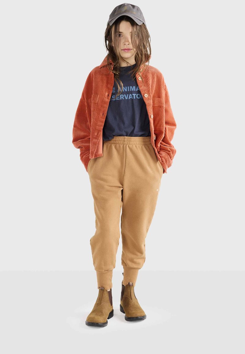 Hnedá kordurová košeľa, námornícke grafické tričko, béžové tepláky a hnedé členkové topánky. Čiapka je šedá s nenápadným logom. Neformálny outfit.