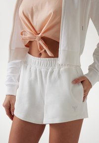 Weiße Baumwoll-Shorts mit elastischem Bund und Seitentaschen, mit einem dreieckigen Logo aus Strass. Darunter ein rosa geknoteter Crop-Top.