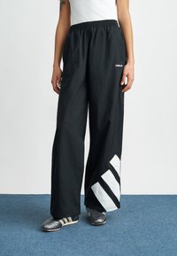 Schwarze leichte Hose mit elastischem Bund, versehen mit großen weißen diagonalen Streifen am unteren Bein und einer glatten Textur.