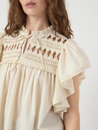 Cream-colored blouse featuring a smocked bodice, tiered ruffle sleeves, and a tie closure at the neckline. Soft texture and loose fit.