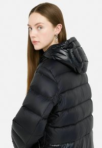 Giubbotto imbottito nero con finitura lucida, design con cappuccio e texture trapuntata. Presenta un collo alto e dettagli di cucitura in evidenza.