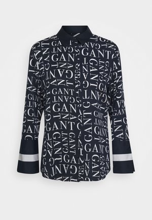 Chemise noire à manches longues avec boutons, ornée du logo GANT blanc en différentes orientations sur toute la surface, col et poignets noirs unis avec une bande blanche sur les poignets.