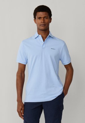 Mand iført lyseblå kortærmet polo med "Hackett"-logo, kombineret med mørkeblå cargobukser, stående foran en ensfarvet baggrund.