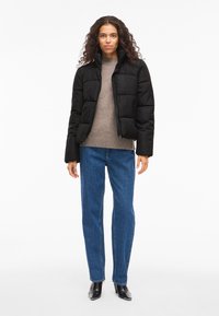 Čierna prešívaná páperová bunda s vysokým golierom a zapínaním na zips, v kombinácii s hnedým pleteným svetrom a modrými džínsami s priamymi nohavicami.