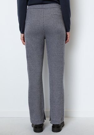 Persoon die grijze geruite rechte broek met achterzakken en zwarte schoenen met dikke zolen draagt, staand tegen een witte muur.