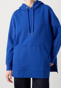 Sudadera azul con un gran bolsillo frontal, capucha con cordón y puños acanalados. Hecha de material suave, presenta un corte relajado y aberturas laterales.