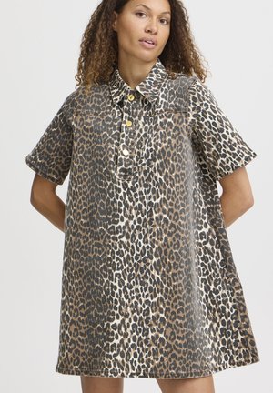Mujer con cabello rizado que lleva un vestido de manga corta, con cuello, estampado de leopardo y botones dorados sobre un fondo claro y sencillo.