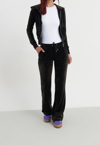 Chaleco de terciopelo negro con cremallera y pantalones a juego con cordón, cuentan con dos bolsillos laterales, combinados con zapatillas moradas sobre un fondo claro.
