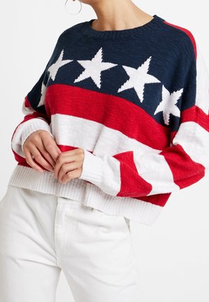 Kobieta ubrana w niebieski, czerwony i biały sweter z białymi gwiazdkami i paskami, do tego białe spodnie i kolczyki koła.