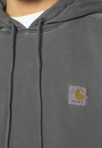 Felpa grigia Carhartt con cordini visibili e patch con logo sul petto, indossata sopra una maglietta bianca a collo rotondo.