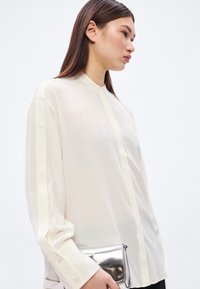 Világos bézs áttetsző blúz gombolással, mandarin gallérral és kontrasztos krémszínű csíkokkal az ujjakon. Fémes borítéktáskával kiegészítve.