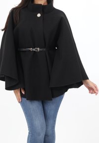 Svart cape i ullblandning med vida ärmar, fäst med en enda metallknapp och ett svart skärp i midjan, kombinerad med blå jeans.