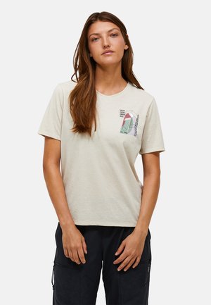 Ung kvinde med langt brunt hår iført en lysebeige grafisk t-shirt og sorte bukser, stående foran en ensfarvet hvid baggrund.