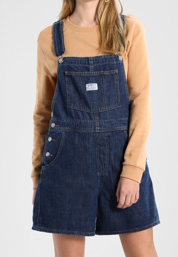 Femme portant une salopette courte en denim bleu foncé avec une poche avant et des détails à boutons, associée à une chemise à manches longues beige.