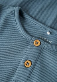 Ribotas mėlynas audinys marškinėliai, turintys apvalų iškirpimą, du oranžiniai mygtukai ir baltai atspausdintas prekės ženklas.