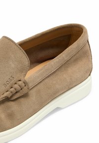 Barna velúr, slip-on cipő fehér gumitalppal, sima felülettel és finom varrásokkal a felső részen. A logó a cipő oldalán van debütálva.