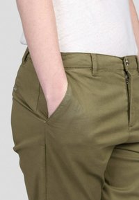 Pantalons vert olive avec poches, fabriqués à partir d'un tissu lisse. Présentent une fermeture éclair et des coutures visibles le long de la taille.
