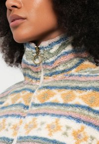 Fliisist lukuga jakk, millel on kõrge krae, pastelltoonides mitmevärvilised triibumustrid ja kuldne tõmblukk.