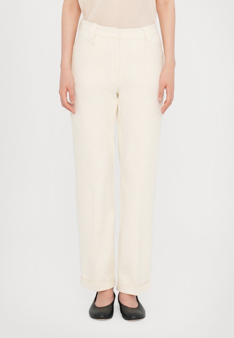 Partie inférieure d'une personne portant un pantalon crème taille haute, coupe droite, avec ourlets retournés et chaussures plates noires sur un fond blanc.