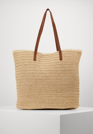 Beige vævet tote bag med to lange brune læder skulderremme, vist stående på en hvid piedestal mod en ensfarvet baggrund.