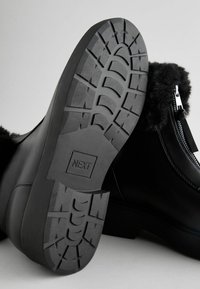 Black ankle boot featuring a textured rubber sole with a tread pattern, synthetic upper, and a zip closure on the side.