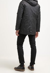 Homme portant une veste à capuche noire, un pantalon noir retroussé, des chaussettes noires et des chaussures en cuir noires avec des semelles brunes, se tenant contre un fond blanc.