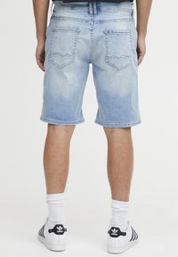 Pantalones cortos de mezclilla azul claro con un corte recto, dos bolsillos traseros con un diseño de costura ondulada y un dobladillo terminado. Llevados con calcetines y zapatos blancos.