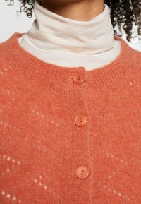 Cardigan tricoté corail avec un motif texturé, présentant trois boutons ronds orange et un col arrondi, porté sur un col roulé blanc.