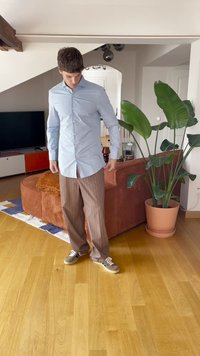 Muž upravující světle modrou košili stojí na dřevěné podlaze poblíž hnědé pohovky a velkého zeleného květináče v moderním obývacím pokoji.