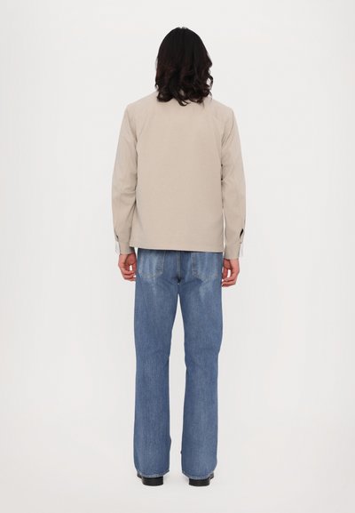 Cămașă bej cu mâneci lungi, cu croi drept și cusături vizibile, asortată cu blugi evazați de un albastru deschis. Design simplu, detalii minime.