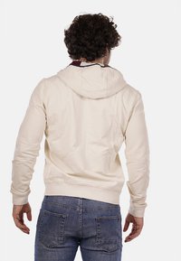 Felpa con cappuccio color crema, dal texture morbido, con polsini e orlo aderenti, caratterizzata da un bordo interno del cappuccio in contrasto. Abbinata a jeans blu.