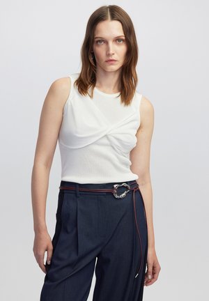 Top bianco senza maniche con design frontale attorcigliato, abbinato a pantaloni blu scuro a vita alta e una cintura decorativa intrecciata.