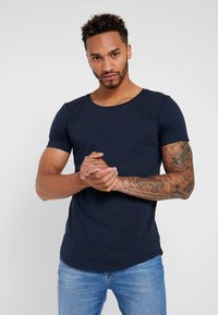 T-shirt blu navy a maniche corte in morbido cotone, con colletto rotondo e orlo curvo. Il modello ha tatuaggi visibili sulle braccia.