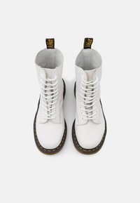 Botas brancas de couro com atacadores, com um cano alto, interior texturado cinza e solas de borracha preta com costura amarela e alças de puxar.