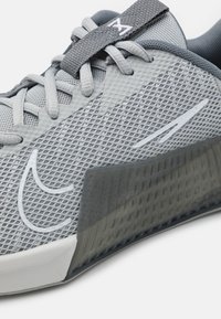 Jasnoszary but sportowy z teksturowaną siateczkową cholewką, wyściełanym kołnierzem, szarymi sznurowadłami i częściowo przezroczystą gumową podeszwą.