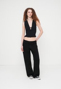 Μαύρο, αμάνικο τοπ με σφηκοφωλιά, βαθιά V-λαιμόκοψη και λεπτομέρεια με κορδόνι, συνδυασμένο με φαρδιά μαύρα παντελόνια και μαύρα αθλητικά παπούτσια.