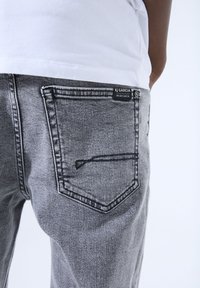 Grijze denim jeans met een lichte verweerde textuur en een achterzak met een geborduurde rand. Merklabel zichtbaar nabij de tailleband.