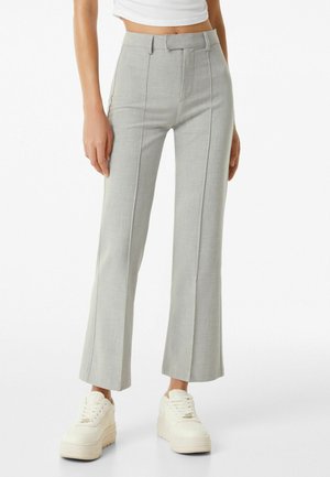 Mujer con pantalones de talle alto de color gris claro, entallados con costuras frontales, y zapatillas blancas con plataforma, posando frente a un fondo blanco.