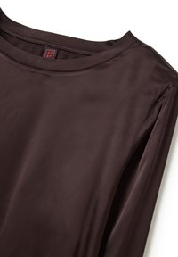 Top de satén marrón oscuro de manga larga con cuello redondo y una pequeña etiqueta de marca roja y negra visible dentro del cuello.