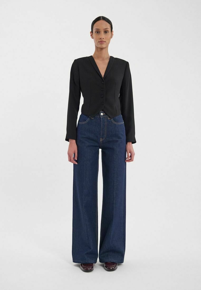 Blazer noir ajusté avec un col en V, cinq boutons et manches longues, associé à un jean en denim foncé à jambes larges et des chaussures marron.