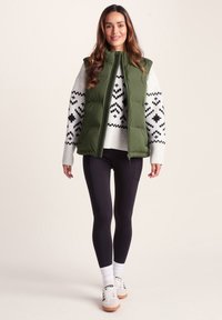 Green puffer vest with zip closure, worn over a white patterned sweater, paired with black leggings and white sneakers.