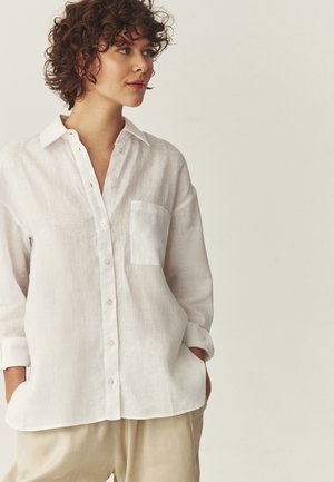 Kobieta o kręconych włosach, ubrana w luźną białą koszulę na guziki z podwiniętymi rękawami oraz beżowe spodnie, stojąca z rękami w kieszeniach.