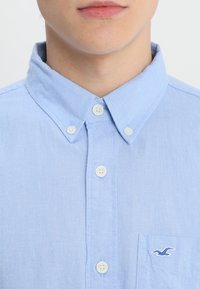 Muž v košili s krátkým rukávem světle modré barvy s límečkem a vyšitým logem racka na kapsičce na hrudi.