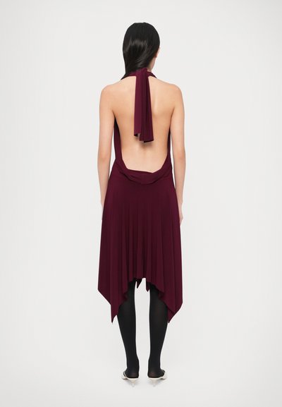Rochie cu halter bordeaux, cu spatele adânc și tiv asimetric, fluid. Material neted și accent drapat în talie. Purtată cu ciorapi negri.