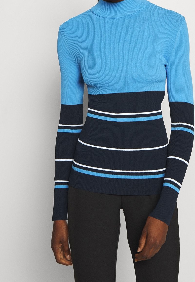 Femme portant un pull en maille côtelée à manches longues bleu et marine avec des rayures horizontales blanches et bleu clair, associé à un pantalon noir.