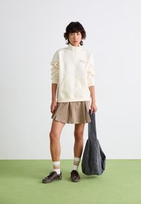 Un sweat-shirt crème avec une fermeture éclair mi-haut, une poche avant et une broderie verte. Associé à une jupe beige plissée, un tote gris et des baskets marron.