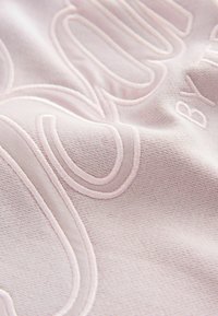 Gaiši rozā audums ar reljefu dizainu, kas izceļ smalku faktūru un diegu apdari. Uzsvars uz paceltu šuvēm un gludu fona.