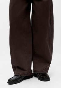 Pantalon large marron foncé porté avec des mocassins noirs en cuir, sur fond blanc.