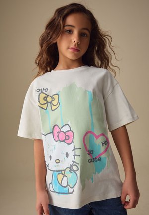 Junges Mädchen mit welligen braunen Haaren trägt ein weißes Hello Kitty T-Shirt, das Schleifen, ein Herz mit dem Text "so cute" sowie hellblaue und grüne Farbspritzer zeigt.